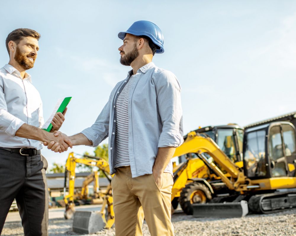 Builder choosing heavy machinery for construction with a sales consultant standing with some documents on the open ground of a shop with special vehicles